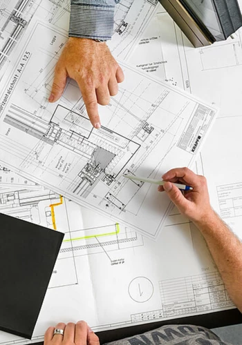 Architects or engineers consulting over detailed blueprints, pointing at a sectional drawing with a pencil during a meeting.